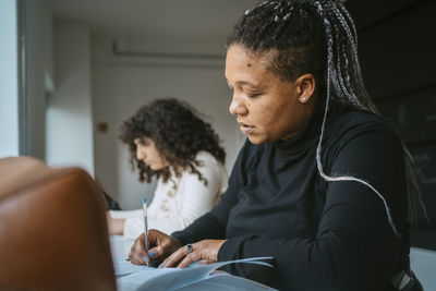 Woman with braided hair writing in book while sitting by friend at university