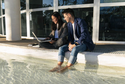 Businessman with feet dipped in pool sitting with colleague using laptop