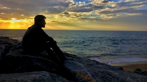 Woman sitting on rock at beach against sky during sunset