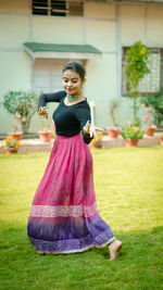 Side view of young woman dancing on grassy field against building in yard