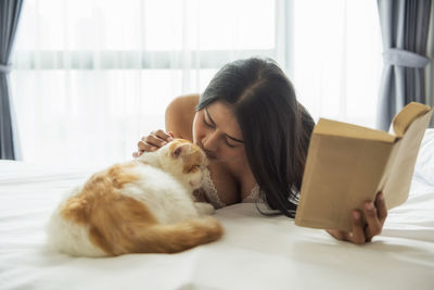 Woman kissing cat while reading book on bed at home