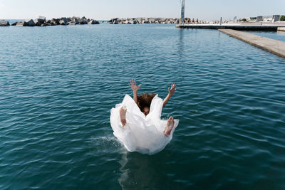 Unrecognizable young barefoot female in white wedding dress falling in rippling lake near pier looking at camera