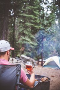 Rear view of man with drink sitting by barbecue in forest