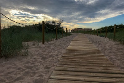 Boardwalk leading towards beach against sky during sunset