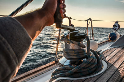 Cropped hand of man rigging rope on boat in sea against sky during sunset