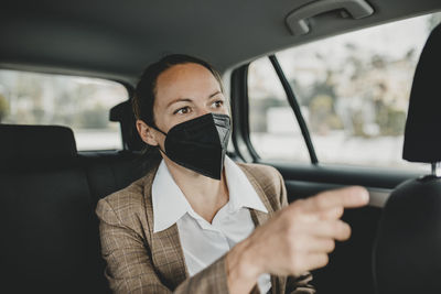 Female professional in face mask gesturing while traveling in taxi during covid-19