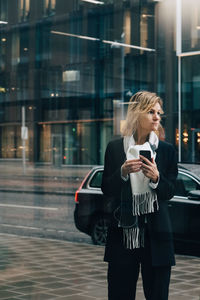Businesswoman with smart phone looking away while standing on sidewalk against building in city