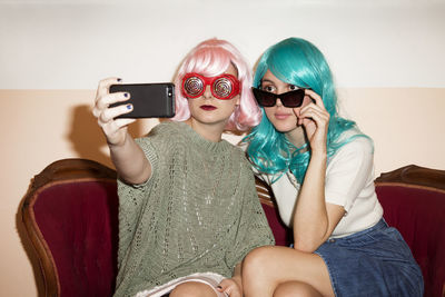 Women wearing wigs clicking selfie while sitting on couch against wall