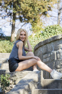 Portrait of young woman sitting on retaining wall against trees