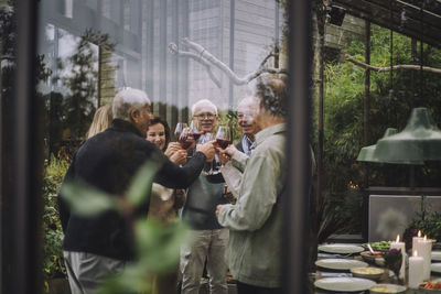 Retired male and female friends toasting drinks at party seen through glass