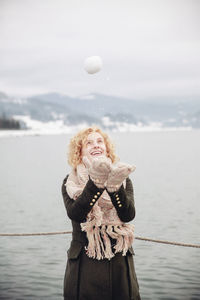 Happy woman playing with snowball by lake against sky