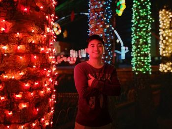 Portrait of young man standing against illuminated tree trunks at night