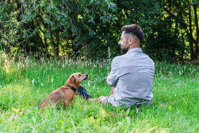 Young european man sitting on grass with his dog in park. concept of human and pet relationship.
