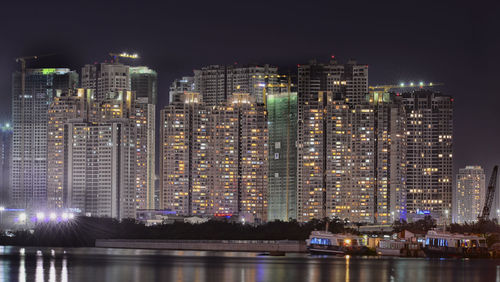 Illuminated modern buildings by river against sky at night