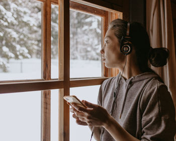 Woman in country house at the window with headphones and mobile phone