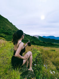 Side view of woman sitting on grassy field against sky