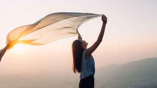 Side view of woman holding sarong against sky during sunset