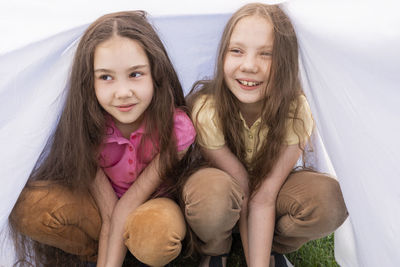 Two cute little sisters with long hair sits under white sheet on grass in meadow. caucasian asian