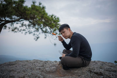 Portrait of young man eating food while sitting on cliff against cloudy sky