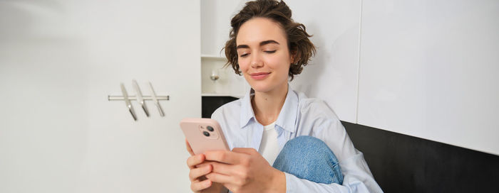 Young woman using mobile phone while standing against wall