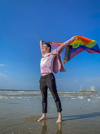 Portrait of a young man standing with a lgbtq flag on the beach against the sky