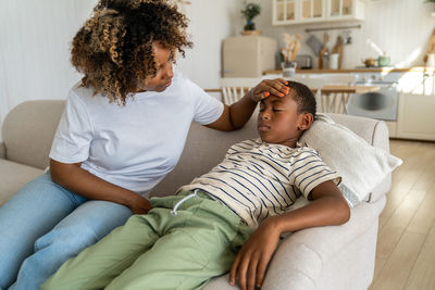 Attentive black woman touch forehead of sick child son sit at home sofa feel unwell weakness malaise