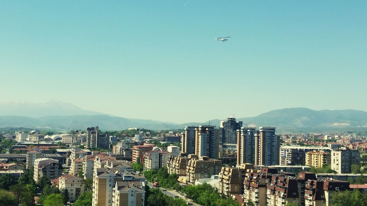 flying, mountain, building exterior, blue, clear sky, city, sky, outdoors, mountain range, cityscape, no people, architecture, airplane, day, urban skyline, nature, drone