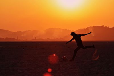 Silhouette man kicking football against sky during sunset