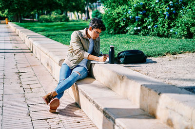 Focused male student sitting on stone bench in university campus and writing in notepad while doing homework on sunny day