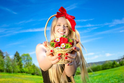 Smiling young woman holding basket of strawberries and daisies at field against sky