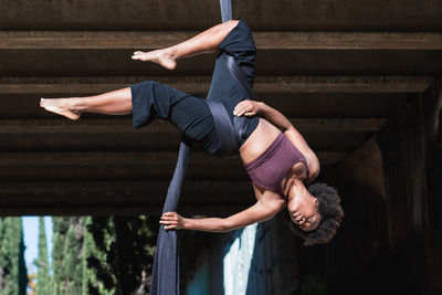 African american female balancing upside down on straps in splits while practicing aerial yoga in city