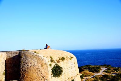 Man standing on rock by sea against clear blue sky