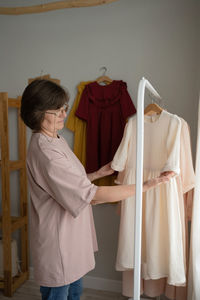 Middle aged woman in casual clothes and glasses examining sleeves of handmade dresses while working in light workshop