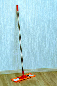 Nickel plated mop with orange handle, near bright wall with rag pad of orange color on floor. 