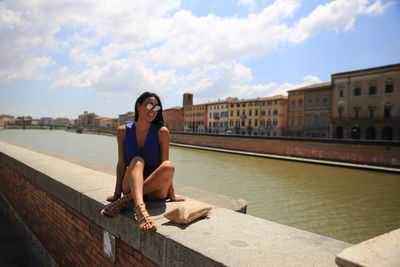 Full length of smiling young woman wearing sunglasses while sitting on retaining wall against sky in city