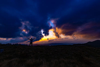 Silhouette man with arms outstretched standing on landscape against sky during sunset
