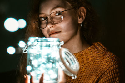 Close-up portrait of young woman holding illuminated string lights in jar
