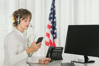  woman customer support call operator in office using mobile phone with blank screen computer