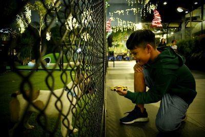 Side view of boy looking through chainlink fence at night