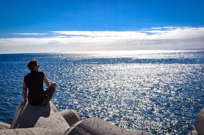 Rear view of man sitting on tetrapod at seashore against blue sky during sunny day