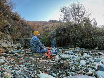 Side view of man sitting on rock by bonfire against sky