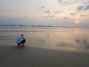 Rear view of man crouching at beach against sky during sunset