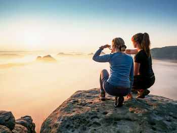 Side view of mother and daughter sitting on rock against sky during sunset