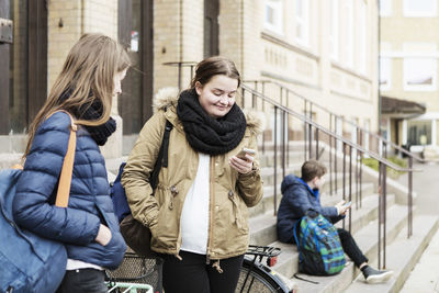 Schoolgirl looking at female friend using mobile phone outside school building
