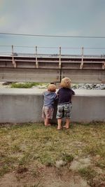Rear view full length of siblings looking at bridge while standing by retaining wall