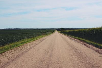 Empty dirt road amidst field against sky