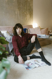Portrait of young man with long hair sitting on floor near sofa at home