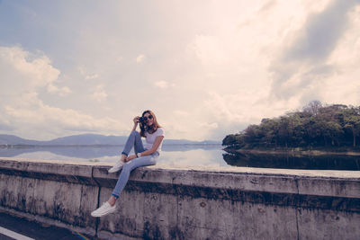 Portrait of woman photographing while sitting against lake and sky