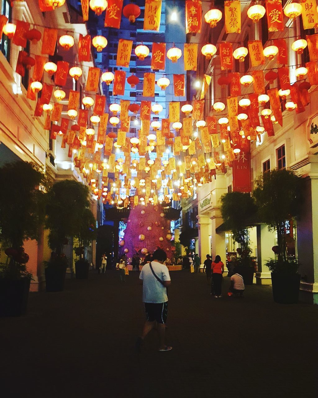 REAR VIEW OF PEOPLE WALKING ON ILLUMINATED STREET AMIDST BUILDINGS