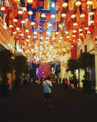 Rear view of people walking on illuminated street amidst buildings at night